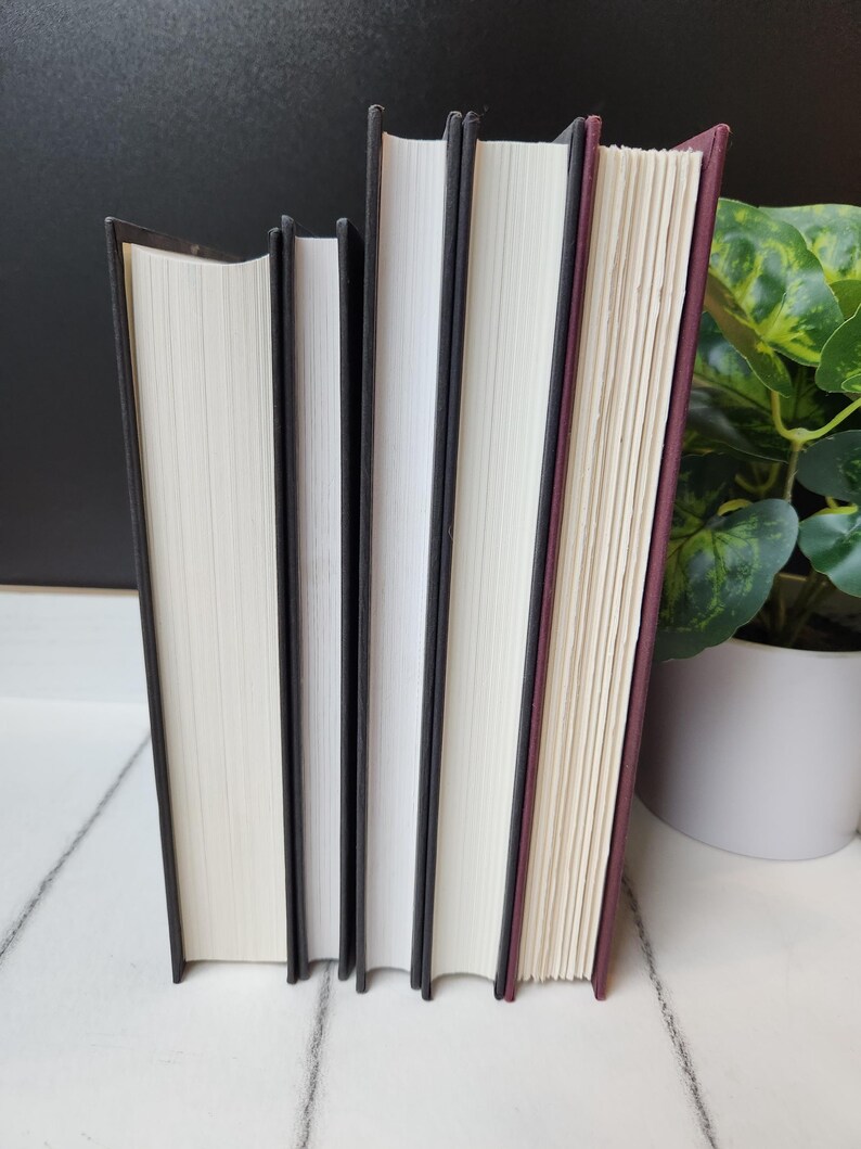 以下が含まれることがあります： 白いページと黒い背表紙を持つハードカバーの本が4冊、白い表面に立てられています。一番右の本は、えんじ色の背表紙です。