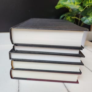 以下が含まれることがあります： 黒い表紙と白いページを持つハードカバーの本が5冊積み重ねられています。本は白い表面にきれいに積み重ねられています。