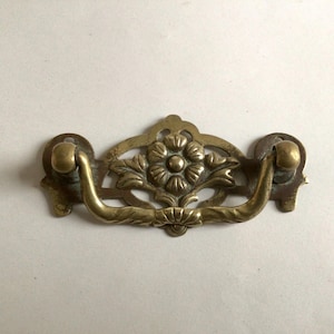 May include: A brass drawer pull with a floral design. The pull has a loop handle and a decorative backplate with a stylised flower in the centre.
