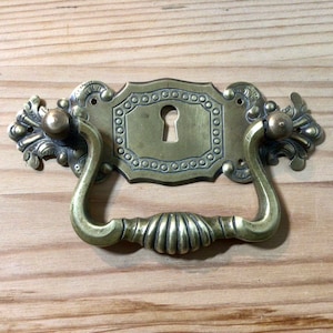 May include: Antique brass drawer pull with a keyhole and ornate design. The pull has a decorative, curved handle and a rectangular plate with a keyhole.