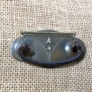May include: Antique brass drawer pull with an oval-shaped plate and a curved handle. The plate has a decorative design with a floral motif.