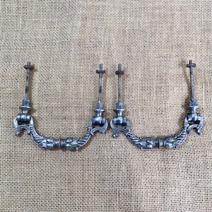 May include: Four antique silver-toned drawer pulls with ornate, curved designs. Each pull features a decorative floral pattern and is attached to a threaded rod with a nut. The pulls are arranged on a textured, neutral-toned surface.