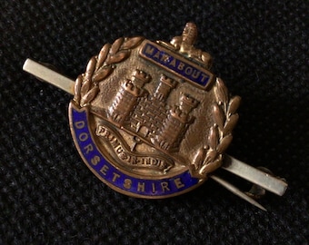 May include: A gold-colored pin with a blue enamel border. The pin features a castle with a sword and the words "Dorsetshire" and "Marabout" on it.