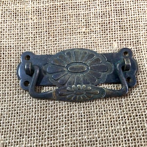 May include: Antique brass drawer pull with a floral design. The pull has a rectangular shape with a curved handle.