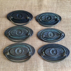 May include: Six antique oval cabinet pulls with a star design in the centre. The pulls are made of brass and have a dark green patina.