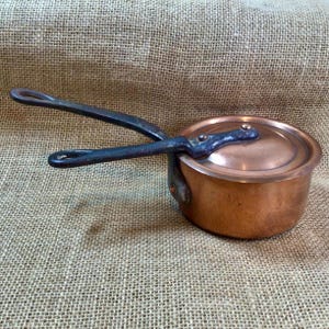 May include: A small copper saucepan with a lid and a black iron handle. The saucepan is sitting on a hessian surface.