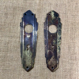 May include: Two antique brass door plates featuring an Art Deco design. The plates have a keyhole and a round opening for a door knob. The plates are slightly tarnished and have a worn patina.