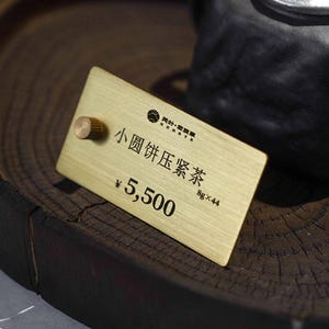 May include: A gold rectangular tag with Chinese text and the number 5,500, placed on a dark wooden tray. A black ceramic teapot with a lid sits in the background. The tray has a dark brown, textured surface.