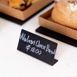 Puede incluir: Un letrero de pizarra negro con texto blanco que dice "Pan de queso y nuez ¥18.00".