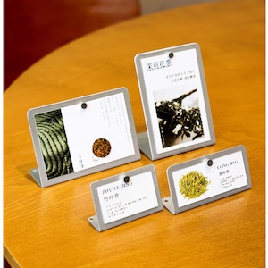 Puede incluir: Cuatro pequeños expositores de metal gris con tarjetas blancas. Las tarjetas presentan imágenes de hojas de té y texto en chino. Los expositores están sobre una mesa de madera. Las hojas de té son verdes y marrones.