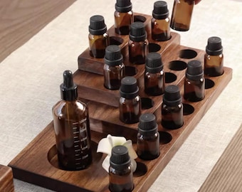 May include: A wooden tiered stand with 18 round holes to hold essential oil bottles. The stand is made of dark brown wood and has a natural finish. There are 17 amber glass bottles with black caps in the holes, and one bottle is being placed in a hole by a hand. There is a larger amber glass bottle with a dropper on the bottom shelf.