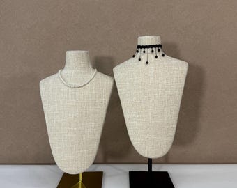 May include: Two jewellery display busts. One bust displays a pearl necklace, the other a black beaded choker. The busts are covered in a beige fabric and mounted on stands. One stand is gold, the other black. The background is a solid brown.