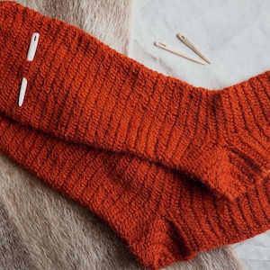 Puede incluir: Un par de calcetines de punto de color óxido con una textura acanalada, sobre una superficie texturizada. Dos agujas de hilo de plástico blanco y dos agujas de madera se colocan cerca de los calcetines. Los calcetines están hechos a mano.