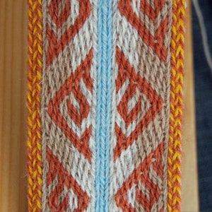 May include: Close-up of a woven textile with a repeating diamond pattern. The design features orange, white, and tan diamonds on a light blue and tan background, with yellow and orange borders. The textile is likely used for decorative purposes.