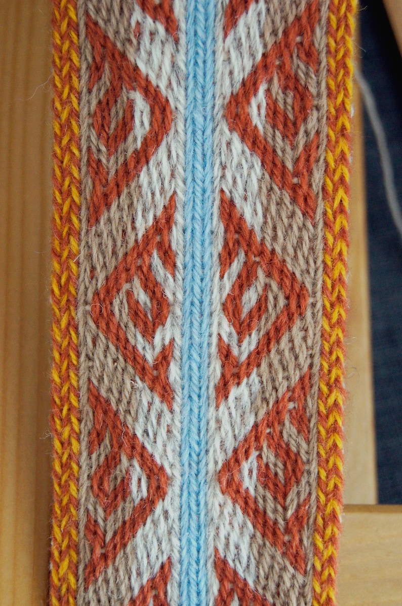 May include: Close-up of a woven textile with a repeating diamond pattern. The design features orange, white, and tan diamonds on a light blue and tan background, with yellow and orange borders. The textile is likely used for decorative purposes.