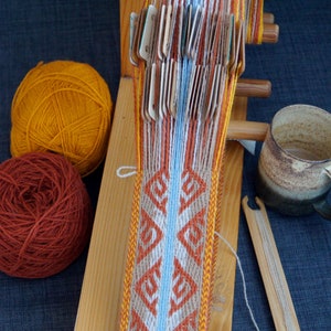 May include: A wooden weaving loom with a partially woven textile featuring geometric patterns in orange, white, and blue. Two balls of yarn, one orange and one rust-colored, sit nearby. A ceramic mug and weaving tools are also present.