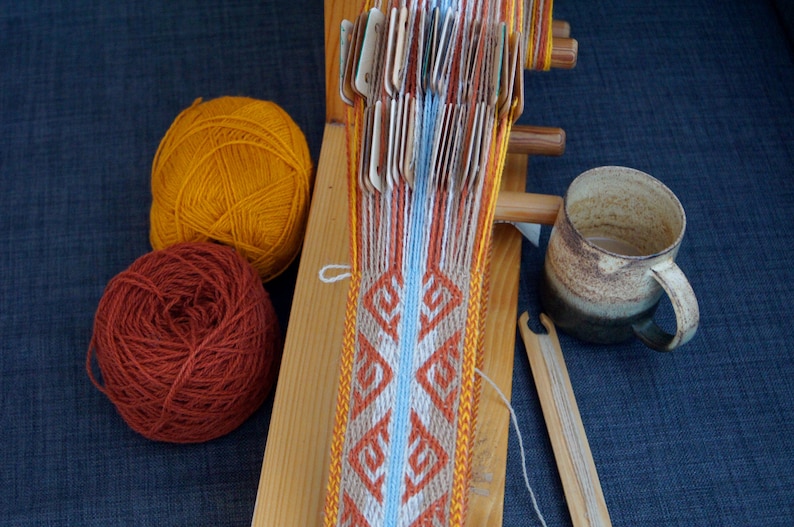 May include: A wooden weaving loom with a partially woven textile featuring geometric patterns in orange, white, and blue. Two balls of yarn, one orange and one rust-colored, sit nearby. A ceramic mug and weaving tools are also present.