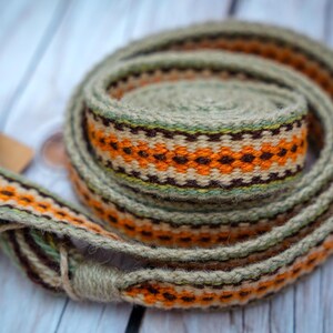 Op de afbeelding: Een geweven riem met een herhalend patroon van oranje, bruine en cr&egrave;mekleurige geometrische vormen. De riem is opgerold en toont het gedetailleerde ontwerp. De achtergrond is een lichtgekleurd houten oppervlak.