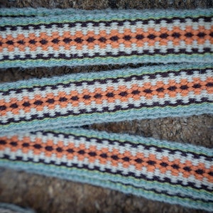 Op de afbeelding: Close-up van geweven lint met een herhalend geometrisch patroon. Het lint heeft een lichtblauwe achtergrond met een patroon van oranje, witte en donkerbruine ruitvormen. Het lint wordt waarschijnlijk gebruikt voor knutselen of decoratieve doeleinden.