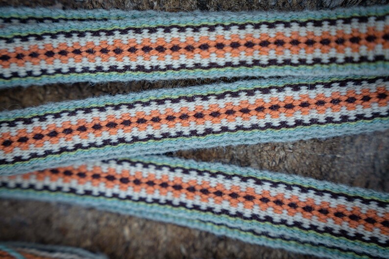 Op de afbeelding: Close-up van geweven lint met een herhalend geometrisch patroon. Het lint heeft een lichtblauwe achtergrond met een patroon van oranje, witte en donkerbruine ruitvormen. Het lint wordt waarschijnlijk gebruikt voor knutselen of decoratieve doeleinden.