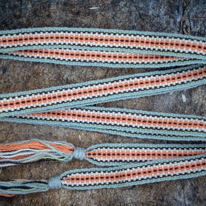 Op de afbeelding: Een geweven riem met een herhalend patroon van oranje, zwarte en witte ruiten op een lichtgroene achtergrond. De riem heeft kwasten aan de uiteinden en wordt gepresenteerd op een houten oppervlak.