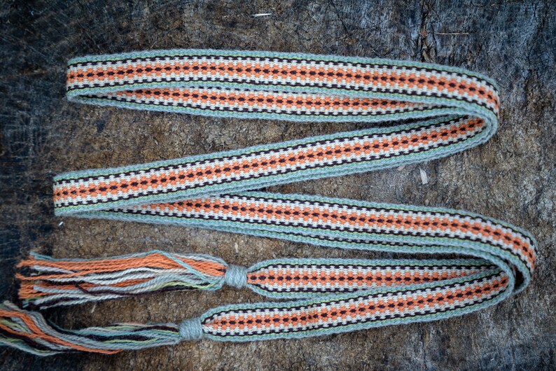 Op de afbeelding: Een geweven riem met een herhalend patroon van oranje, zwarte en witte ruiten op een lichtgroene achtergrond. De riem heeft kwasten aan de uiteinden en wordt gepresenteerd op een houten oppervlak.