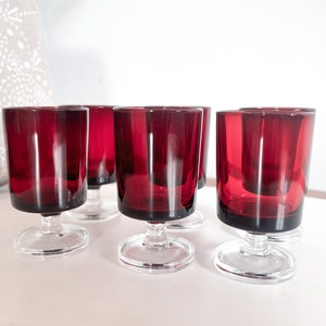 Puede incluir: Seis vasos de chupito de vidrio transparente con base roja y tallo transparente. Los vasos están dispuestos en fila sobre una superficie blanca.