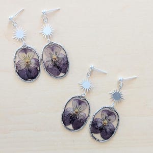 May include: Silver-toned dangle earrings featuring pressed purple pansies encased in oval frames. Each earring has a sunburst accent and a post for pierced ears. The earrings are displayed on a light wooden surface.