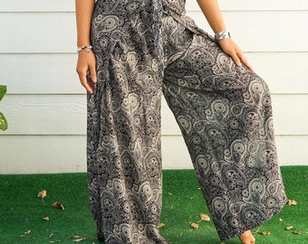 May include: A woman wearing a white tank top and black and white paisley print pants. She is smiling and has long brown hair. She is standing in front of a white wall with a window behind her.