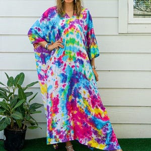 Op de afbeelding: Een enkellange kaftan-jurk met een levendig tie-dye patroon in tinten felroze, elektrisch blauw, zonnig geel en paars. Het kledingstuk heeft een los, vloeiend silhouet met wijde mouwen en een eenvoudig ontwerp met een V-hals.