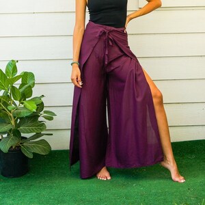 May include: Plum-colored palazzo pants with a high slit, tied at the waist. The pants are paired with a black tank top. The model is standing on green turf in front of a white clapboard wall.