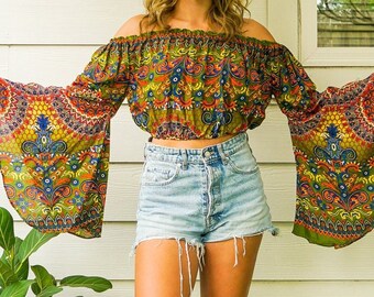 May include: A woman wearing a green, blue, and orange patterned off-the-shoulder top with bell sleeves. The top is cropped and has a gathered neckline. She is also wearing denim shorts.