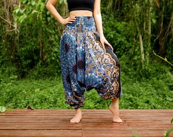 Puede incluir: Una mujer con un top negro sin mangas y pantalones de harén con estampado azul y marrón. Los pantalones tienen una pierna ancha y una cintura con cordón. La mujer está de pie en una plataforma de madera con vegetación verde en el fondo.