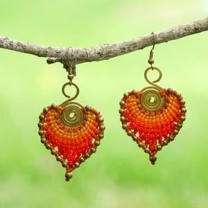 Puede incluir: Un par de pendientes hechos a mano con forma de corazón. Los pendientes están elaborados con hilo naranja y amarillo, acentuados con pequeñas cuentas doradas. Cuelgan de una rama sobre un fondo verde borroso.