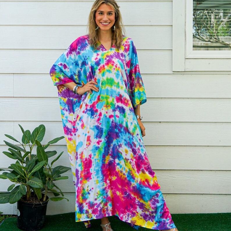 Op de afbeelding: Een enkellange kaftan-jurk met een levendig tie-dye patroon in tinten felroze, elektrisch blauw, zonnig geel en paars. Het kledingstuk heeft een los, vloeiend silhouet met wijde mouwen en een eenvoudig ontwerp met een V-hals.