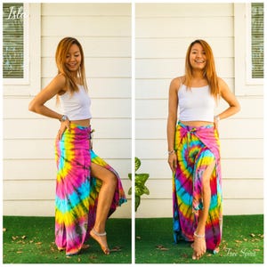 May include: A white sleeveless crop top paired with a long, high-slit skirt featuring a vibrant tie-dye pattern in shades of bright pink, yellow, and turquoise blue. The skirt has a wrap-style front and a tie closure at the waist.