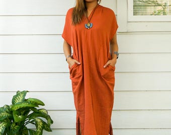 Puede incluir: Un vestido midi de color óxido con mangas cortas, escote en V y dos bolsillos delanteros grandes. El vestido presenta aberturas laterales en el dobladillo y un acabado de tela texturizada. Se combina con un sombrero beige de ala ancha, un collar azul y sandalias.