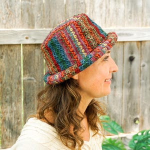 Op de afbeelding: Een patroonhoed met een rand, met een levendige mix van kleuren, waaronder rood, blauw, groen en geel. De stof heeft een geweven textuur. De hoed is een stijlvol accessoire.