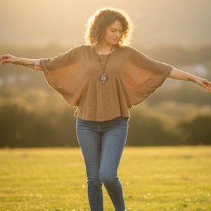 May include: A woman in a brown, flowing top with wide sleeves and blue jeans. The top has a round neckline and a pendant necklace. She is standing in a grassy field with her arms outstretched, bathed in sunlight.