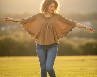 May include: A woman wearing a brown, loose-fitting top with wide sleeves and blue jeans. The top has a round neckline and a pendant necklace. The woman is standing in a grassy field with her arms outstretched.
