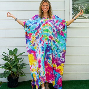 Op de afbeelding: Een vloerlange kaftan met een levendig tie-dye patroon in tinten felroze, geel, turquoise en paars. Het kledingstuk heeft een los, vloeiend silhouet met wijde mouwen, een V-hals en een witte basiskleur die door de gehele stof heen zichtbaar is.