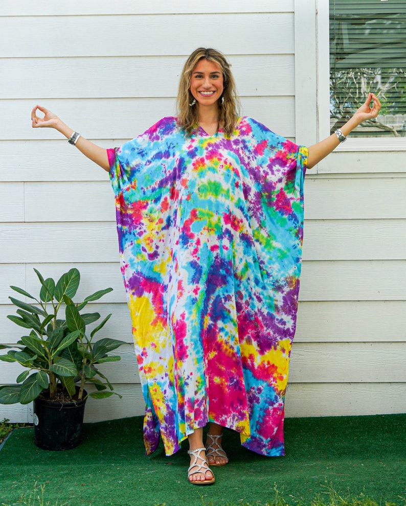 Op de afbeelding: Een vloerlange kaftan met een levendig tie-dye patroon in tinten felroze, geel, turquoise en paars. Het kledingstuk heeft een los, vloeiend silhouet met wijde mouwen, een V-hals en een witte basiskleur die door de gehele stof heen zichtbaar is.