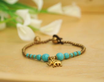 May include: A gold-colored bracelet featuring turquoise beads and a small gold elephant charm. The bracelet is strung on a woven cord and has a clasp closure. The background is a light wooden surface with white flowers.