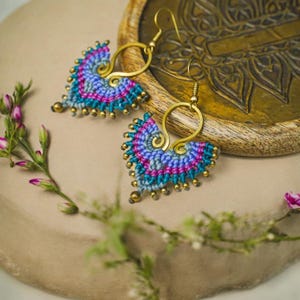 May include: A pair of handmade earrings with a heart-shaped design. The earrings are made with colorful macrame and have gold-colored accents.