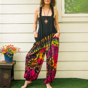 May include: A woman wearing a black tie-dye jumpsuit with a colorful pattern. The jumpsuit has a wide leg and a loose fit. The woman is standing in front of a white wall with a window behind her.