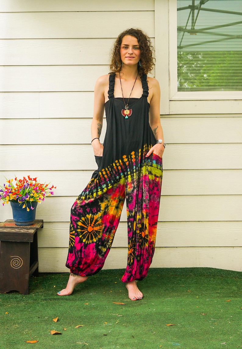 May include: A woman wearing a black tie-dye jumpsuit with a colorful pattern. The jumpsuit has a wide leg and a loose fit. The woman is standing in front of a white wall with a window behind her.