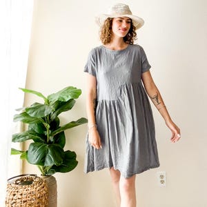 May include: A light gray, short-sleeved dress with a gathered waist and a round neckline. The dress is paired with a straw hat. A potted plant is visible in the background.