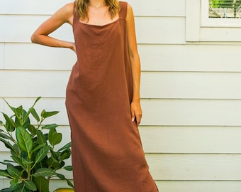 May include: A woman wearing a long, brown, sleeveless dress with a square neckline. She is standing in front of a white wall with a window and green grass.