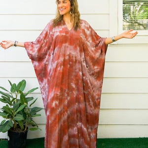 Op de afbeelding: Een enkellange kaftan-jurk met een tie-dye patroon in tinten terracotta, roest en wit. Het kledingstuk heeft een V-hals, wijde vloeiende mouwen en een los, ontspannen silhouet. De tekst Isla en Terracotta Sky zijn zichtbaar in de afbeelding.