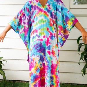 Op de afbeelding: Een enkellange kaftan met een levendig tie-dye patroon in tinten van felroze, elektrisch blauw, zonnig geel en dieppaars. Het kledingstuk heeft een V-hals, wijde mouwen en een losse, ontspannen pasvorm die soepel naar beneden valt.